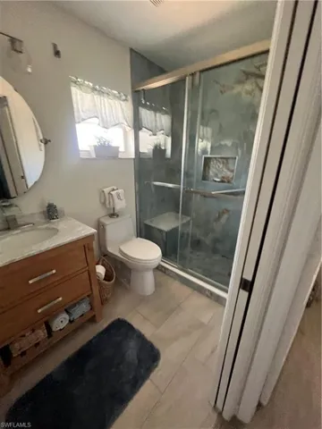 Walk-in shower with glass enclosure, wood-finish vanity with stone countertop, round mirror, window with privacy curtain, and light-toned tile flooring