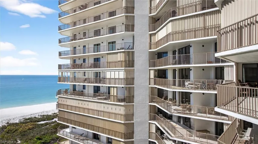 Multi-story building featuring numerous balconies with metal railings, sliding glass doors, and a beachfront location with white sand