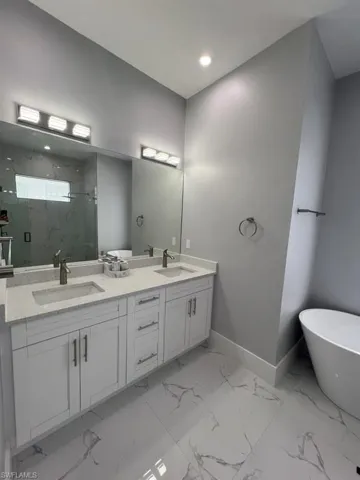 Bathroom featuring a dual vanity with white cabinetry and light-toned countertops, large format marble-look tile flooring, a freestanding soaking tub, and an enclosed shower with matching marble-look tiling