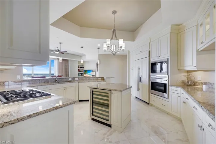 Gourmet kitchen featuring white cabinetry, granite countertops, professional-grade gas cooktop, stainless steel appliances, a central island with wine refrigerator, recessed and pendant lighting, and polished stone flooring throughout