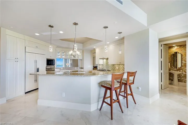 Gourmet kitchen featuring a large waterfall-edge island with seating for two, stainless steel appliances, white cabinetry, pendant lighting, and polished stone flooring throughout the open living area
