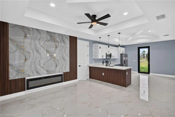 Open-concept living space featuring a textured accent wall with an integrated electric fireplace, a modern kitchen island with pendant lighting, white shaker-style cabinetry, and large format porcelain tile flooring throughout