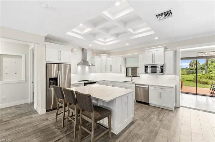 Kitchen featuring appliances with stainless steel finishes, beam ceiling, coffered ceiling, wall chimney range hood, and white cabinets