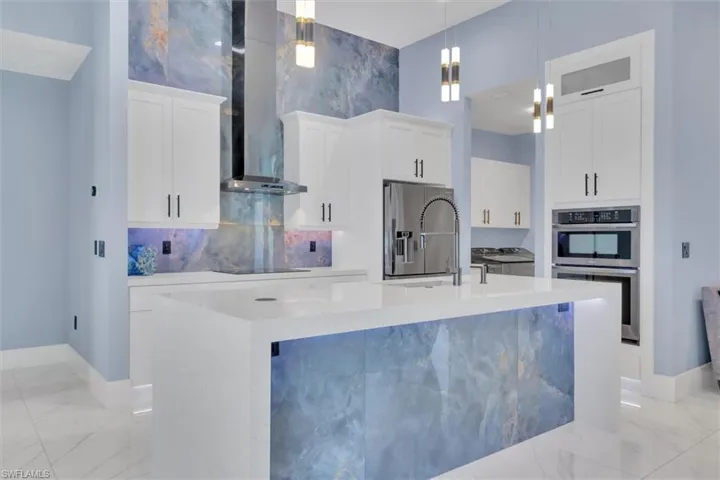 Kitchen with a sink, stainless steel appliances, wall chimney range hood, light marble finish flooring, and a center island with sink