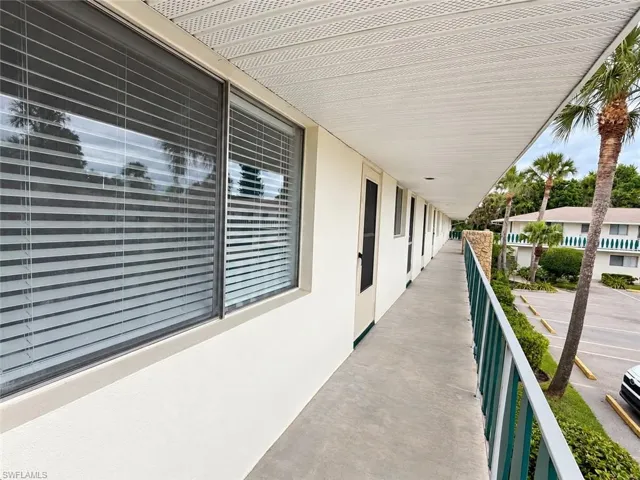 Covered exterior walkway with a concrete surface, featuring a railing, multiple windows with blinds, and exterior doors