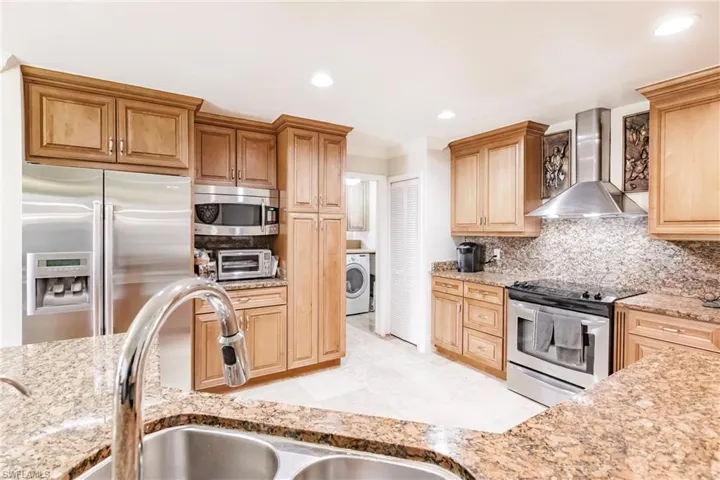 Kitchen with wall chimney range hood, stainless steel appliances, washer / dryer, light stone countertops, and recessed lighting