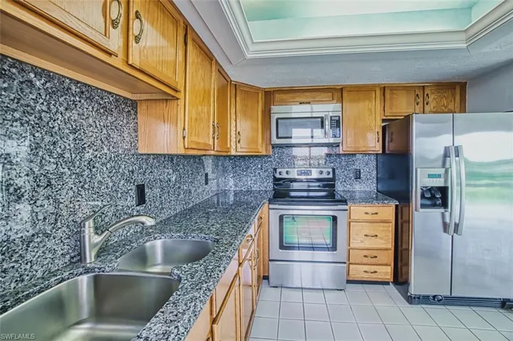 Kitchen with appliances with stainless steel finishes, light tile patterned flooring, brown cabinets, backsplash, and dark stone countertops