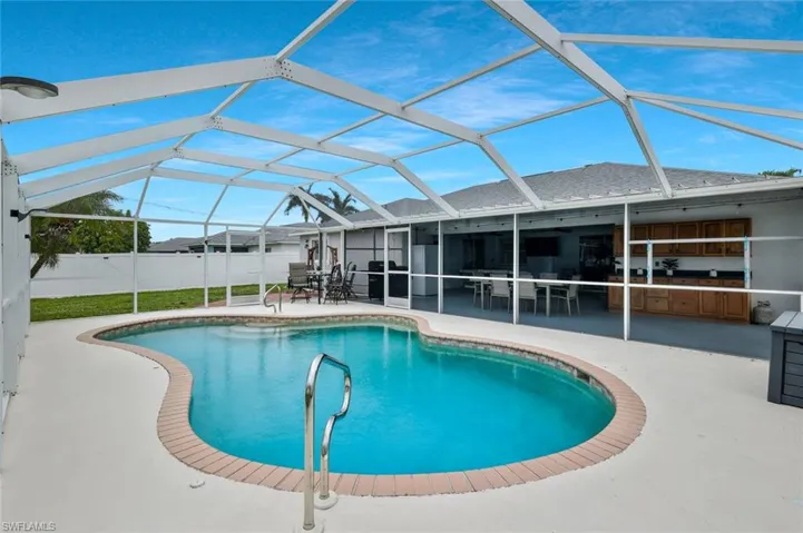 Screened pool enclosure featuring a freeform swimming pool with brick coping, an expansive concrete pool deck, and a covered lanai with built-in cabinetry