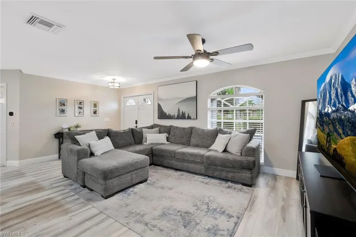 Spacious living area featuring light wood-finish flooring, a contemporary ceiling fan, elegant crown molding, and an arched window with horizontal blinds