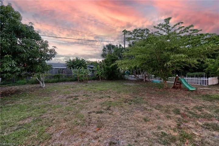 Spacious backyard featuring mature trees, a privacy fence, a white picket fence, and a green slide