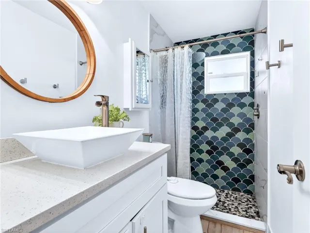 Contemporary bathroom featuring a vessel sink with a bronze-finish faucet, a wood-framed mirror, white cabinetry, and a shower with scallop-patterned tile, a privacy window, and pebble-finish flooring