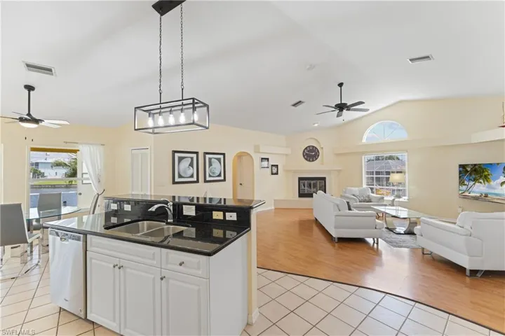 Open-concept living space featuring a kitchen island with dual sinks and dark countertops, tile flooring transitioning to wood-finish flooring, a living area with a fireplace and vaulted ceilings, and a window with an arched transom