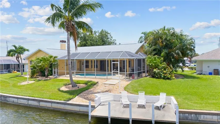 Waterfront property featuring a private boat dock with three white lounge chairs, an expansive screened-in pool enclosure, and a broad green lawn