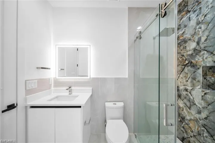 Modern bathroom featuring a single vanity with a white countertop, an illuminated mirror, and a glass-enclosed shower with a rainfall showerhead and patterned wall tile