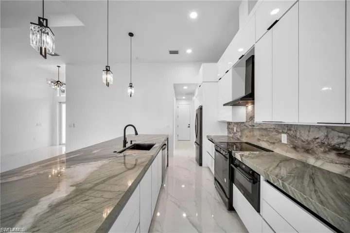 Modern kitchen featuring white high-gloss cabinetry, black appliances, a large center island with an undermount sink, and stone countertops extending to a full-height backsplash