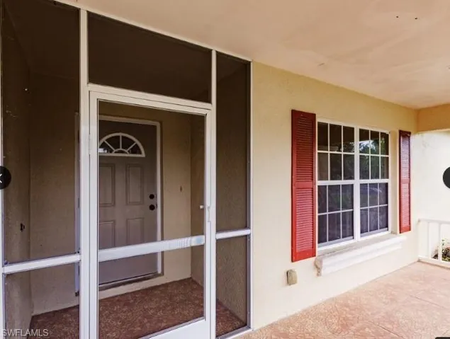 Screened-in porch entry featuring a decorative front door with an arched transom, a large window with red louvered shutters, and a textured concrete floor