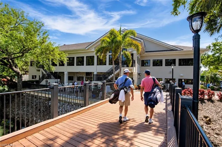 Pedestrian bridge featuring wood-finish decking and metal railings leading to a multi-story clubhouse with a paved patio and mature landscaping