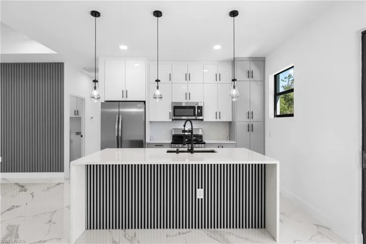 Kitchen with stainless steel appliances, light stone countertops, a kitchen island with sink, white cabinets, and decorative light fixtures