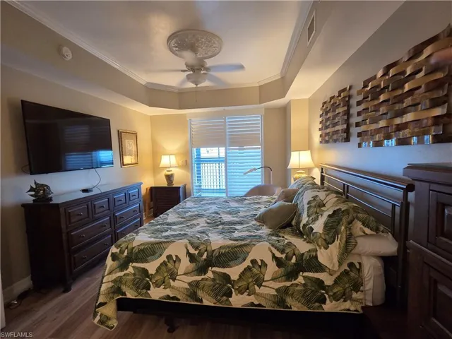Bedroom featuring wood-finish flooring, a tray ceiling with a decorative ceiling medallion and fan, a window with horizontal blinds, and a mounted flat-screen television