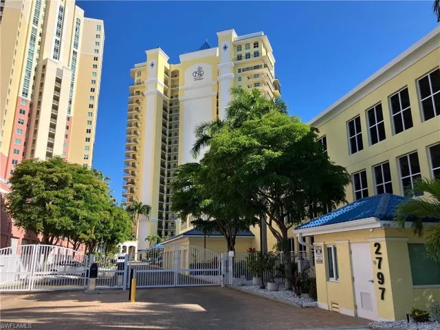 Gated entry to a multi-story building featuring numerous private balconies, mature landscaping, a security kiosk with a blue tile roof, and a paved brick entrance