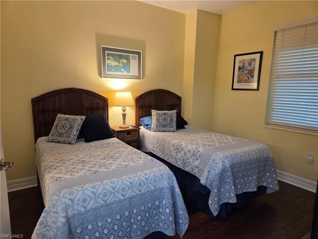 Bedroom featuring two beds with wicker headboards, wood-finish flooring, light yellow painted walls, white trim, and a window with blinds