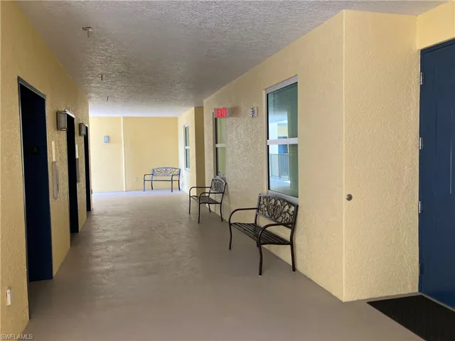 Covered exterior walkway with seating by the elevator just outside your entry door.  Features textured yellow walls and a neutral-toned floor.