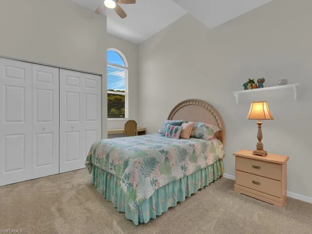 Carpeted room featuring a vaulted ceiling with a ceiling fan, a large arched window with blinds, bi-fold closet doors, and a built-in desktop surface