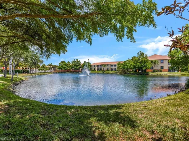 Lakeside setting featuring a water fountain, a pedestrian bridge, lush green landscaping, and multi-story buildings with terracotta-style roof tiles