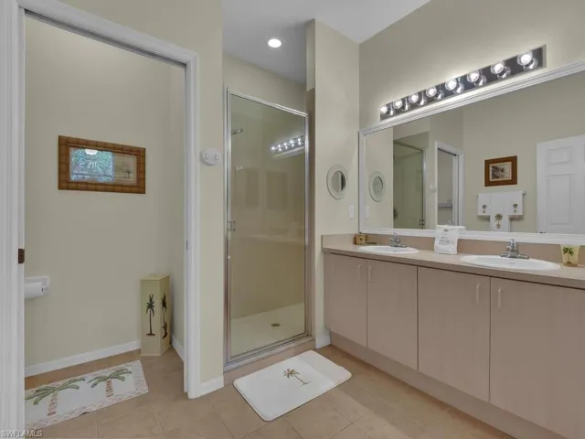 Bathroom featuring a double vanity with integrated sinks, a large mirror with strip lighting, a glass-enclosed shower, and neutral-toned floor tiling