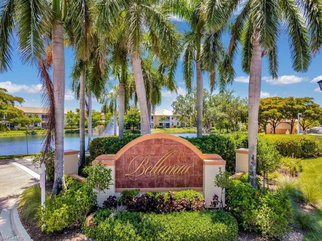 Gated community entrance featuring a decorative arched sign, lush landscaping, mature palm trees, and a prominent water feature with a fountain