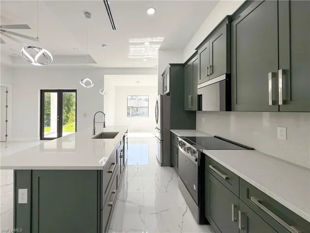 Kitchen featuring green cabinetry, appliances with stainless steel finishes, decorative light fixtures, light stone counters, and a center island with sink