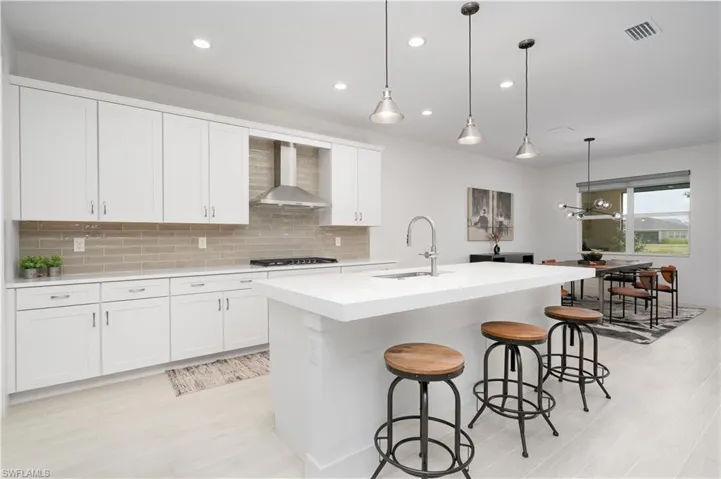 Kitchen with decorative backsplash, decorative light fixtures, an island with sink, wall chimney exhaust hood, and white cabinetry