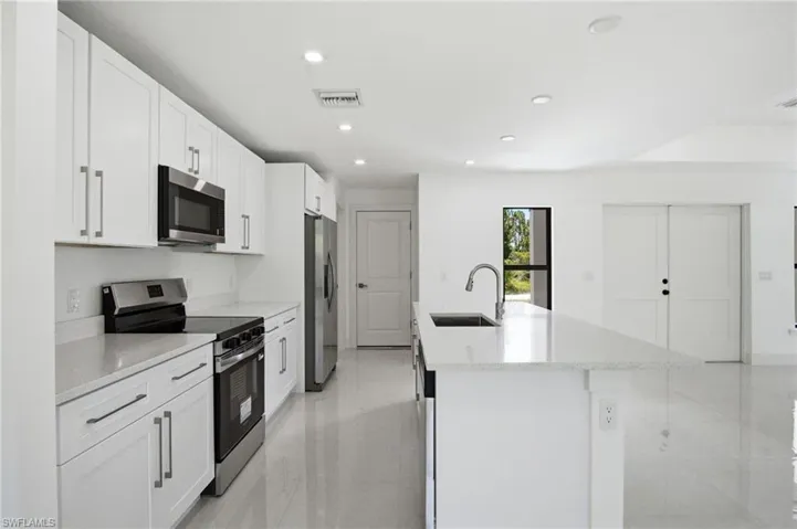 Modern kitchen featuring white cabinetry with brushed nickel hardware, stainless steel appliances, a center island with a sink and gooseneck faucet, light-toned countertops, and recessed lighting
