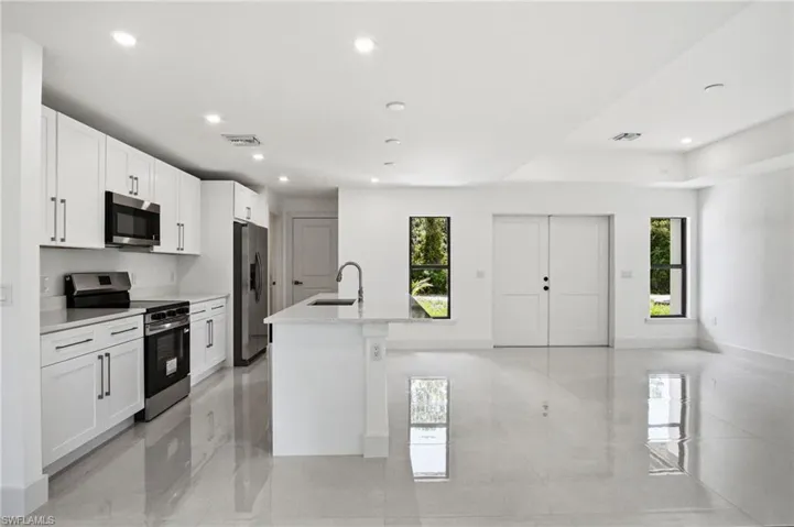 Open-concept kitchen and living area featuring sleek white cabinetry, stainless steel appliances, a center island with an integrated sink, recessed lighting, and large format tile flooring