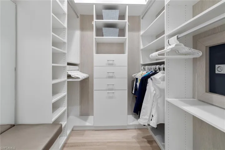 Walk-in closet #2, custom closet system featuring white built-in shelving, integrated drawers with brushed nickel pulls, multiple hanging rods, a built-in upholstered bench, and light wood-finish flooring