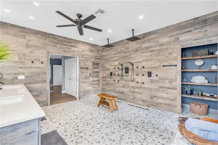 Wood-finish tile walls, pebble-style flooring, dual ceiling-mounted rain shower heads, built-in shelving, and a double vanity with integrated sinks - Virtually Edited Image