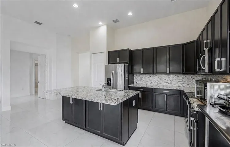 Kitchen with dark cabinetry, light stone counters, stainless steel appliances, a kitchen island with sink, and backsplash
