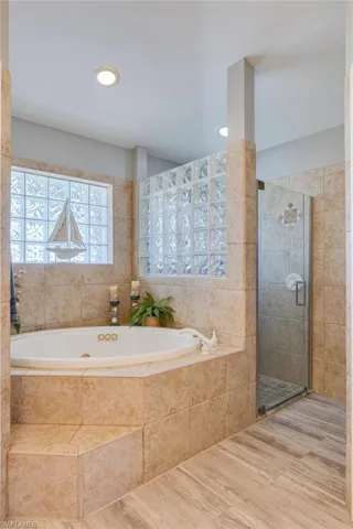 Soaking tub with a tiled surround, glass block window, glass-enclosed shower, wood-finish flooring, and recessed lighting