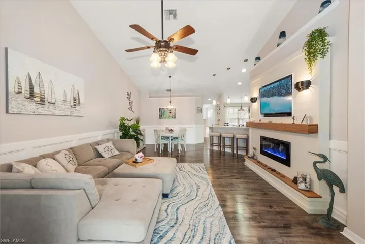 Living area featuring a glass covered fireplace, vaulted ceiling, dark wood-type flooring, a ceiling fan, and a decorative wall