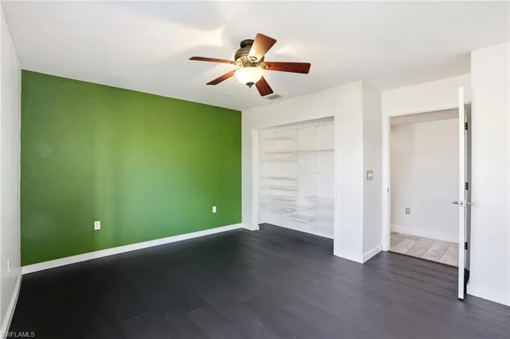Spacious room featuring dark wood-finish flooring, a vibrant accent wall, built-in shelving, a ceiling fan with integrated lighting, and white baseboards