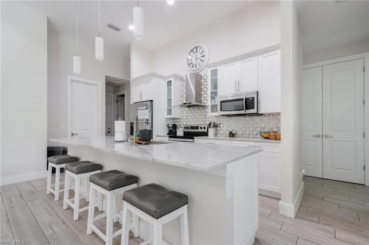 Kitchen with white cabinetry, wall chimney exhaust hood, appliances with stainless steel finishes, decorative light fixtures, and a center island with sink