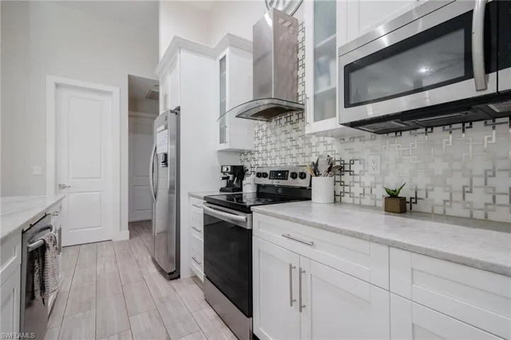 Kitchen with stainless steel appliances, wall chimney range hood, light stone counters, backsplash, and white cabinetry