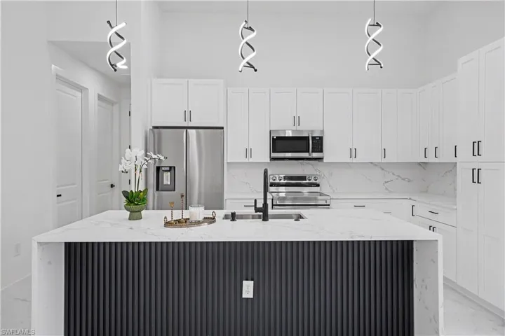 Contemporary kitchen featuring white shaker-style cabinetry, quartz countertops, a waterfall-edge island with vertical ribbed paneling in charcoal finish, stainless steel appliances including a side-by-side refrigerator and built-in microwave, and pendant lighting with modern geometric fixtures suspended above the workspace