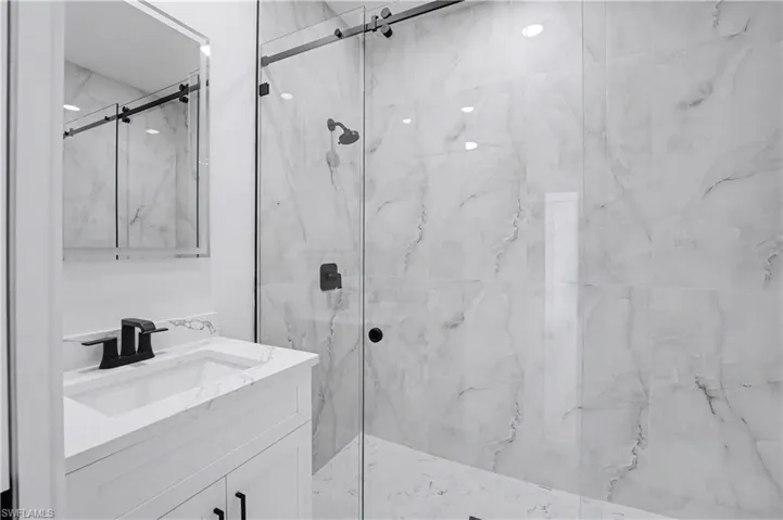 Modern bathroom featuring a floating vanity with white quartz countertop and matte black faucet, large framed mirror with integrated lighting, and a walk-in shower enclosed by frameless glass doors with marble-look tile walls and rainfall showerhead