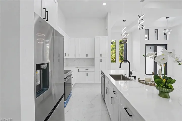 Modern galley kitchen featuring white shaker cabinetry, stainless steel appliances including a French-door refrigerator and built-in oven, white quartz countertops with waterfall edge, matte black faucet with pull-down sprayer, pendant lighting, and light-filled layout with natural light from adjoining windows