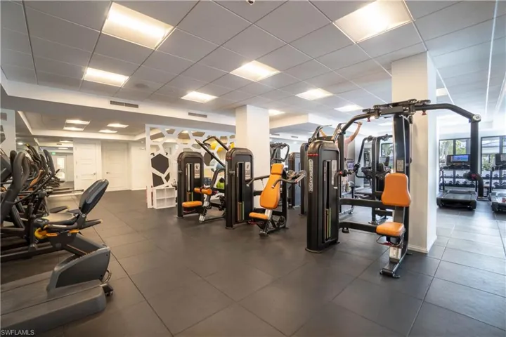 Fitness center with rubberized flooring, abundant overhead lighting, mirrored walls, and a variety of exercise equipment including cardio machines and weight stations
