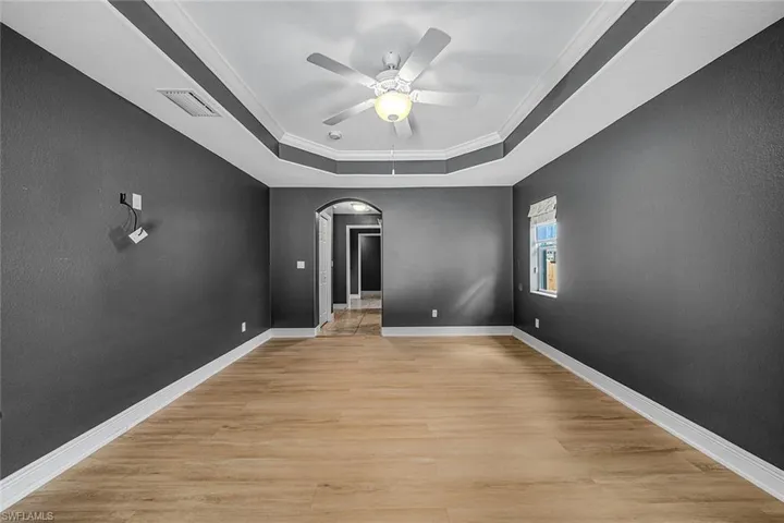 Empty room with arched walkways, crown molding, light wood-style flooring, ceiling fan, and a tray ceiling