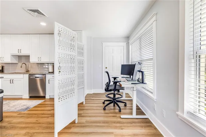Bright interior space with wood-finish flooring, multiple windows with blinds, a white paneled door, and a partial view of a kitchen featuring white cabinetry and stainless steel appliances