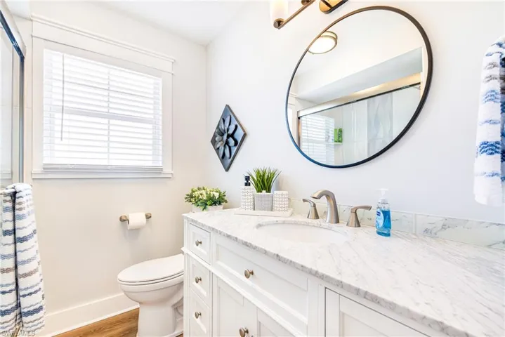 Bathroom featuring a white vanity with paneling and brass hardware, a light-toned stone countertop with an undermount sink, and a circular black-framed mirror