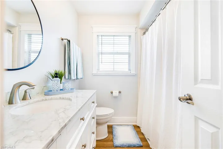 Bathroom featuring a white vanity with a built-in sink and stone countertop, a circular mirror, brushed nickel fixtures, a window with horizontal blinds, and wood-finish flooring